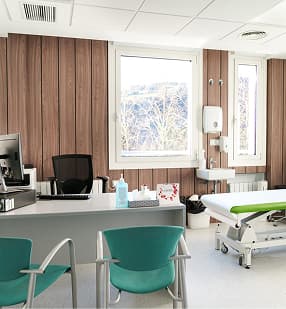 Modern physiotherapy consultation room with wood-paneled walls, treatment bed, desk with teal chairs, and large windows